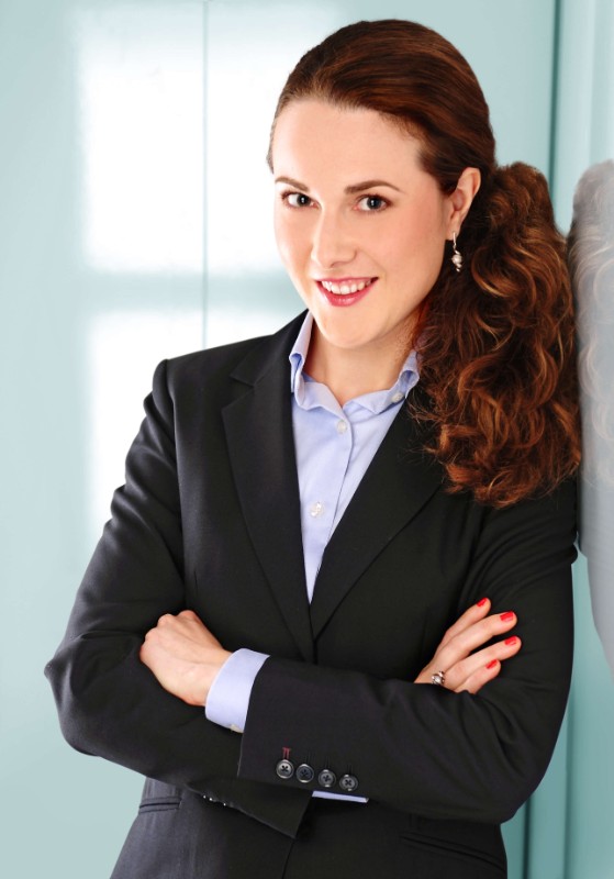 Portrait einer netten jungen Frau mit langen welligen braunen Haaren und dunklem Jackett, die vor hellblauem Hintergrund lacht